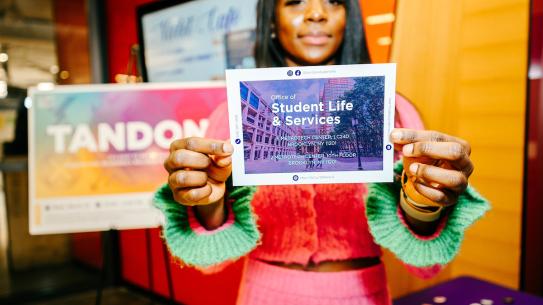 A student in colorful outfit holding a "student Life & Services" postcard 