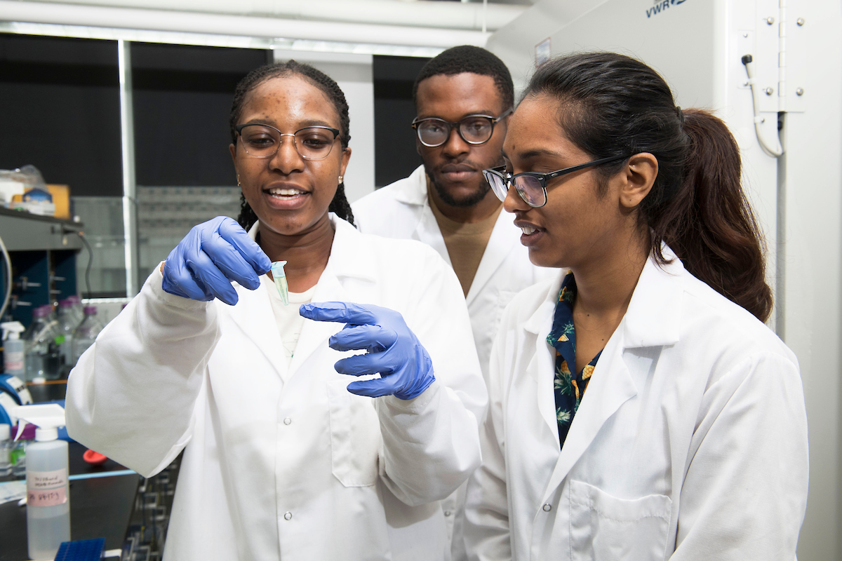 three diverse students in lab coats, one holding a solution in gloved hands