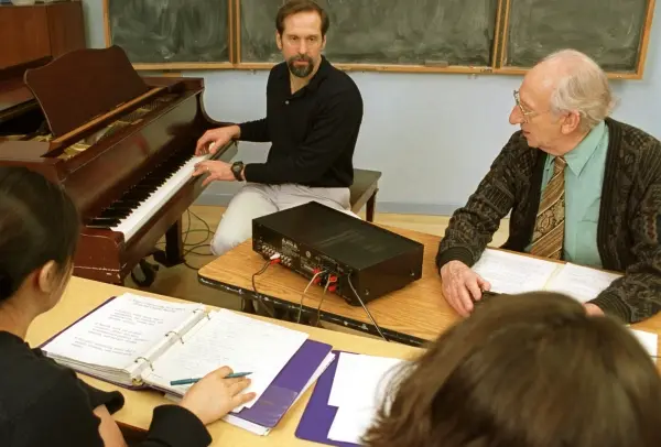 Four individuals are visible. In the center, a man with a beard sits at an upright piano, an older man wearing a patterned cardigan and tie sits at the desk, looking back toward the pianist.