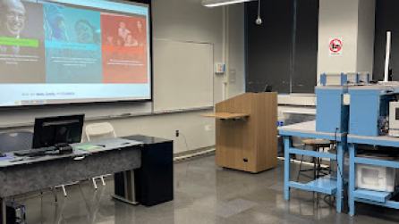 Picture of the JAB 678 teaching space. There is a lectern and a projector screen, as well as a table with a PC.