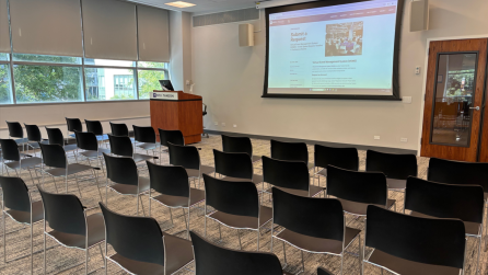 View from the back of the room, with three rows of ten chairs each facing a lectern and a projector screen.