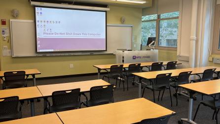 In the foreground, four tables with four chairs at each table. The tables face a whiteboard, a lectern, and a projector screen.
