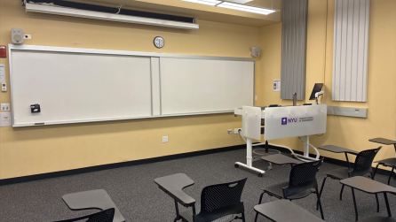 In the foreground, two rows of chairs with tablet arms. The chairs face a whiteboard, a lectern, and a projector screen.