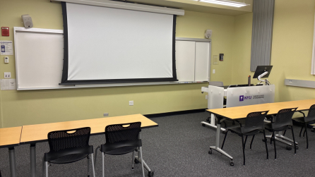 In the foreground, three tables with chairs. The tables face a whiteboard, a lectern, and a projector screen.