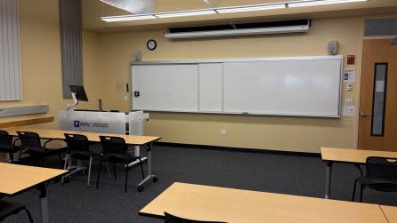 In the foreground, four tables with four chairs at each table. The tables face a whiteboard, a lectern, and a projector screen.