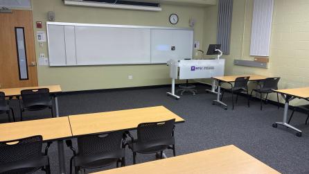 In the foreground, three tables with four chairs at each table on the left side of the room, and two tables with two chairs at each table on the right side of the room. The tables face a whiteboard, a lectern, and a projector screen.