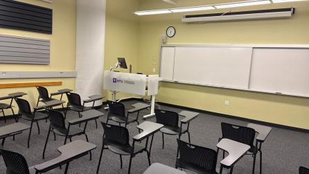 In the foreground, three rows of chairs with tablet arms. The chairs face a whiteboard, a lectern, and a projector screen.