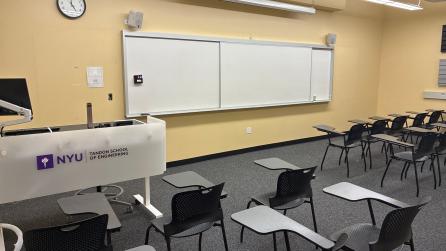 In the foreground, three rows of chairs with tablet arms. The chairs face a whiteboard, a lectern, and a projector screen.