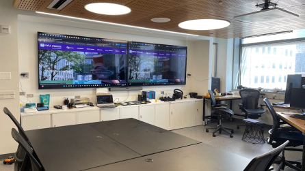 A room with various tables, chairs, and desks with computers. On one wall there are two large displays.