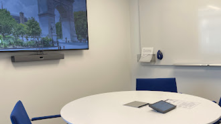 A small room with a round table with four chairs, a whiteboard, and a display on the wall.