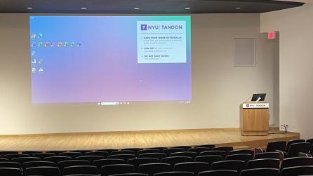 A room with auditorium-style seating, a stage, a podium, and a projector displayed on the wall behind the stage.