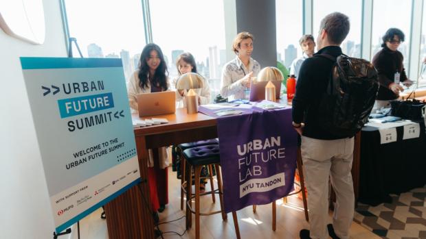 People stand at tables, with a sign in the forefront reading "Urban Future Summit""