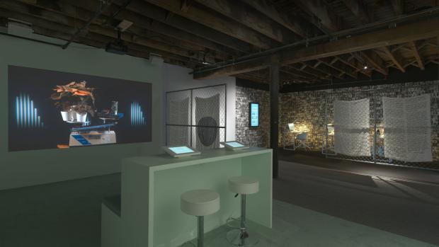 Room with a counter and two stools in the center, facing a wall with an projected image of a scale with plants on one side and various electronics on the other, on it