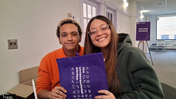 Two students, smiling, holding a bag that reads "The Unconventional Kitchen"