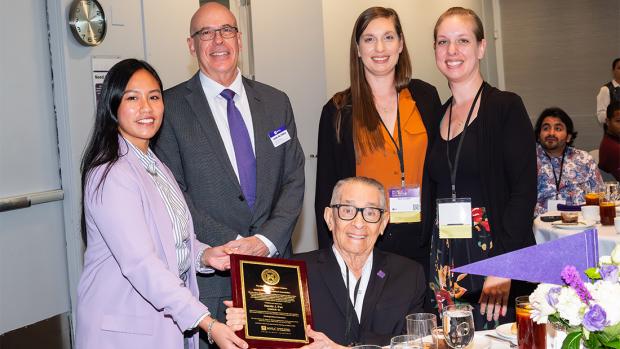 Harold, holding a plaque and seated centered at a table, with four people posed standing around him