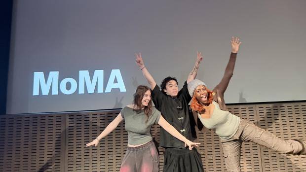 Three students pose on a stage, in front of a screen that reads "NYU Hackathon March 27th, 2026 MoMA"