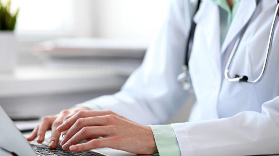 Person in a lab coat and a stethoscope typing on a laptop