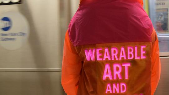 person from the back wearing a vibrant orange and pink jacket. features glowing neon pink text that reads 'WEARABLE ART AND DESIGN'