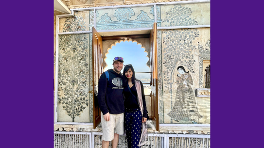 Regvina Oliveira and her husband stand in front of an arched window on wall decorated with decals of patterns, plants and a woman
