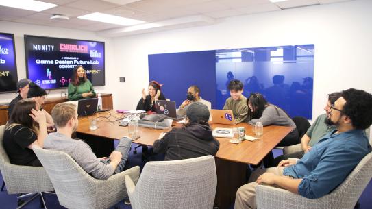 Mitu Khandaker is standing up at the head of the table with a smile on her face addressing NYC GDFL's first cohort.