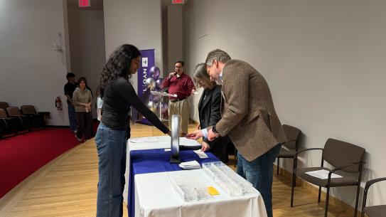 Student to the left standing at a table with her arm outstretched, as the professor on the other side of the table put the engineerings ring on her finger. 