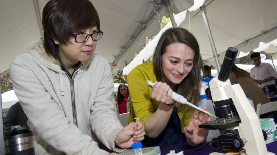 a female student using a pipette at a microscope with a male student observing