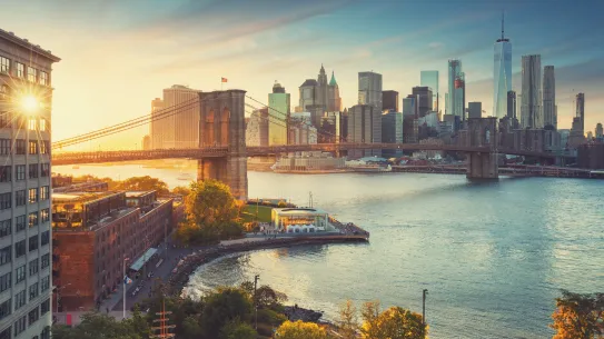 Elevated view of the Brooklyn Bridge stretching across the East River toward the Lower Manhattan skyline at golden hour, with sunlight flaring behind a waterfront building.