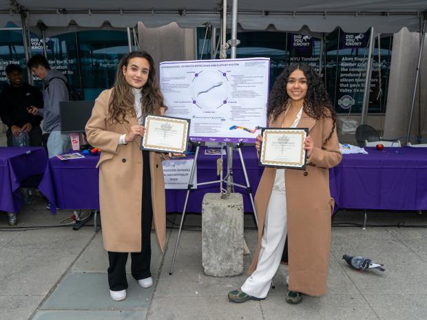 2 women students holding certificates next to large research poster
