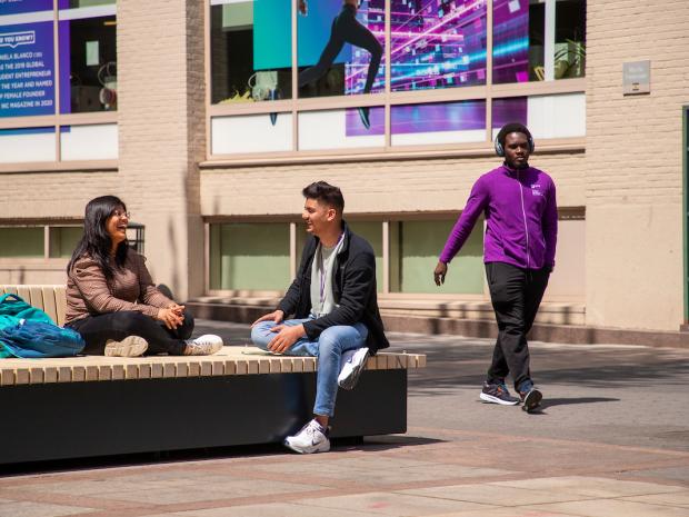 a laughing student on bench with friend while another walks by in NYU gear