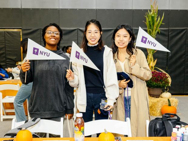 three smiling students at Fall Fest event holding NYU pennants 