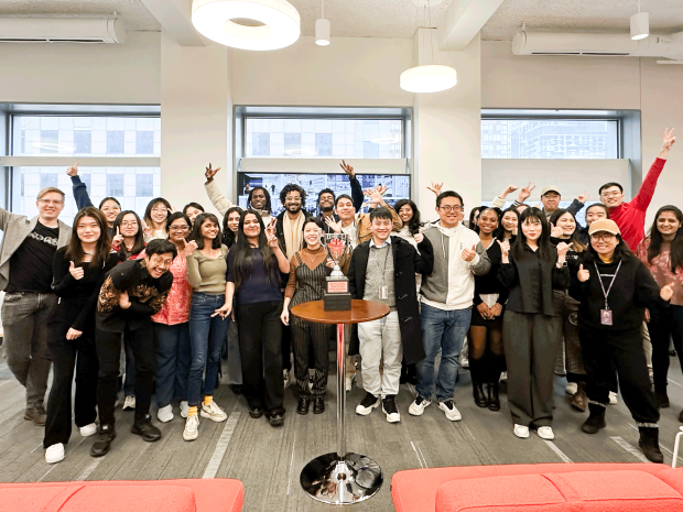 A group of around 30 students and faculty smiling with their arms outstretched around a table with a silver trophy on top.