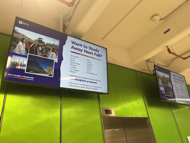 Two screens mounted above the elevators on the first floor of 6 MetroTech. The screens display the same image, which has information about studying abroad in the fall.