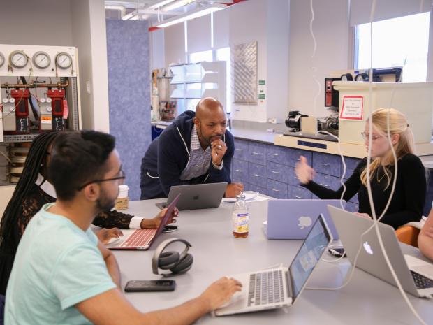 professor leaning over lab table with several students working on laptops