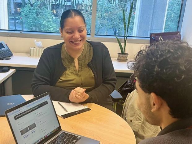 Two people sitting, facing each other, at a table with an open laptop and a notepad.