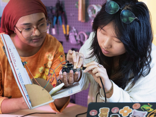 Two students collaborate on an electronics project in a makerspace, examining a wired circuit board and handmade prototype at a worktable with a laptop and engineering tools, highlighting hands-on learning, creativity, and teamwork.