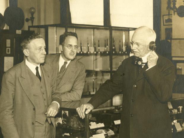 sepia toned photo of men in lab, one holding old telephone to ear