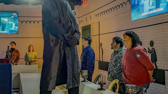 The game host standing on a table while PhD students gather around, getting ready for the next Squid Game challenge.