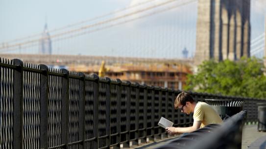 a young man reading on a park bench with the Brooklyn Bridge in the background