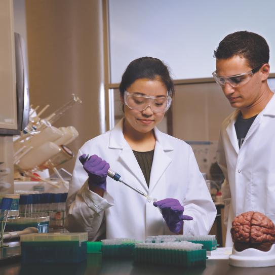 Two students in a lab, wearing lab attire using a mechanical pipette