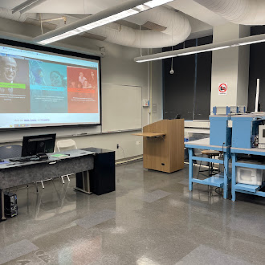 Picture of the JAB 678 teaching space. There is a lectern and a projector screen, as well as a table with a PC.