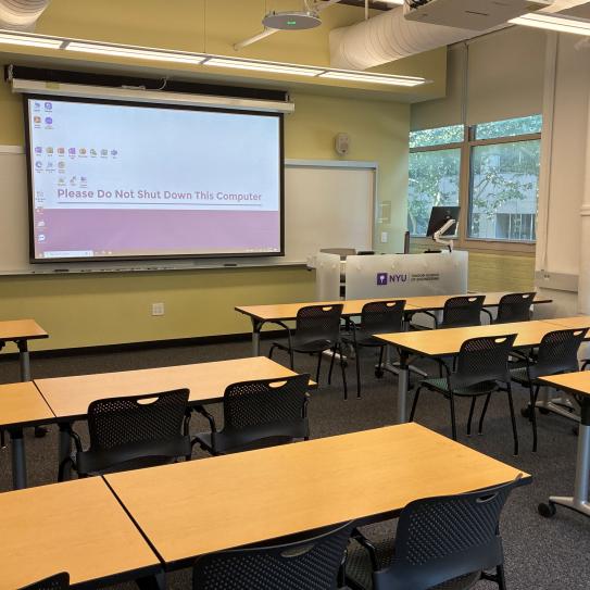In the foreground, four tables with four chairs at each table. The tables face a whiteboard, a lectern, and a projector screen.