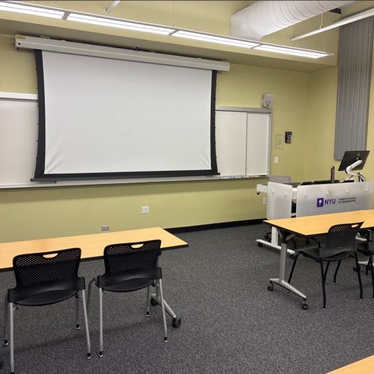 In the foreground, three tables with chairs. The tables face a whiteboard, a lectern, and a projector screen.