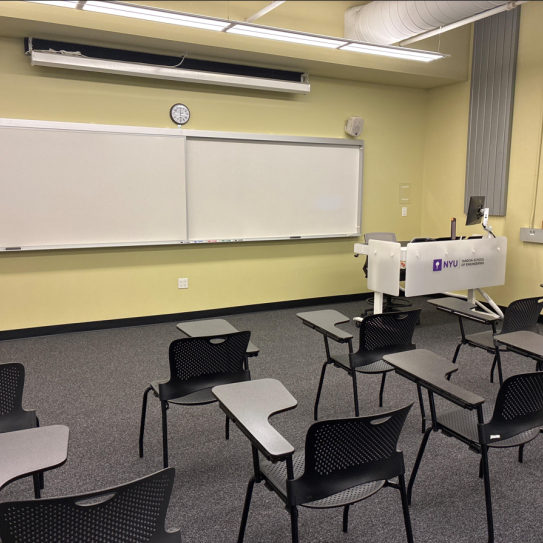 In the foreground, two rows of chairs with tablet arms. The chairs face a whiteboard, a lectern, and a projector screen.