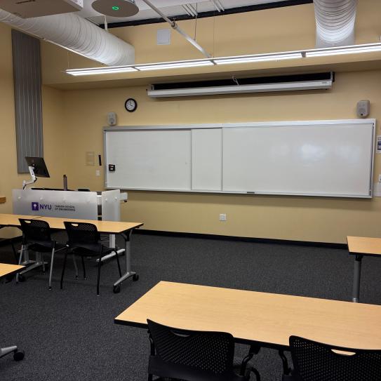 In the foreground, four tables with four chairs at each table. The tables face a whiteboard, a lectern, and a projector screen.