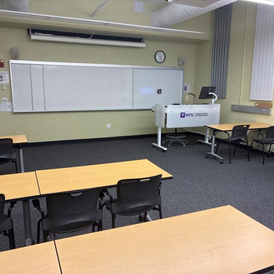 In the foreground, three tables with four chairs at each table on the left side of the room, and two tables with two chairs at each table on the right side of the room. The tables face a whiteboard, a lectern, and a projector screen.