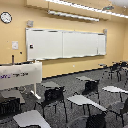 In the foreground, three rows of chairs with tablet arms. The chairs face a whiteboard, a lectern, and a projector screen.