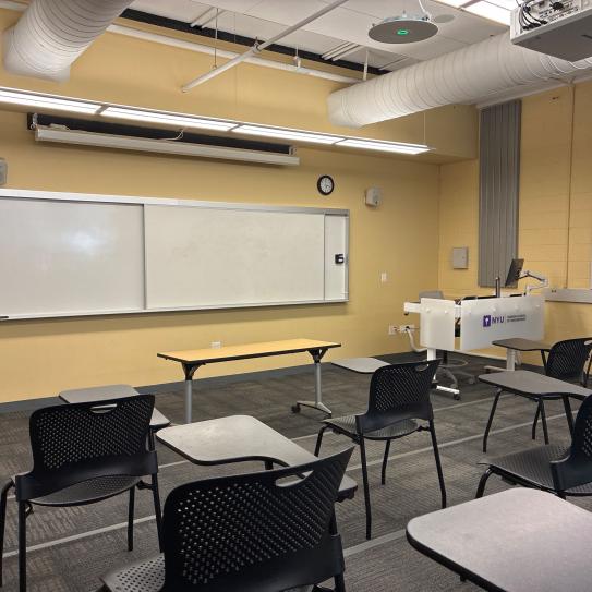 In the foreground, three rows of chairs with tablet arms. The chairs face a whiteboard, a lectern, a table, and a projector screen.
