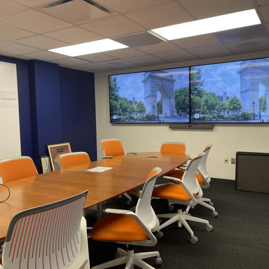 A room with a conference table with 10 chairs. At one end of the table there are two large displays.