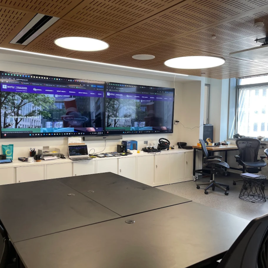 A room with various tables, chairs, and desks with computers. On one wall there are two large displays.