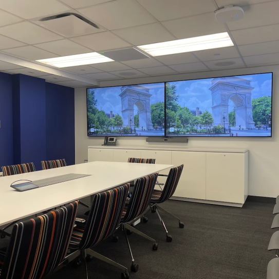 A room with a conference table with 10 chairs. At one end of the table there are two large displays.